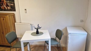 Una habitación pequeña y limpia con una mesa blanca, dos sillas, un radiador, un refrigerador blanco y un cuadro en la pared.