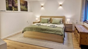 Un dormitorio con una cama, dos mesitas de noche, una mesita auxiliar de madera, una ventana con cortinas e iluminación en la pared.