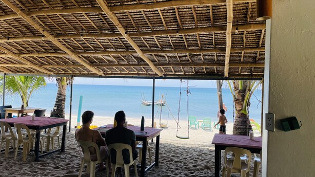 Zona exterior con mesas y sillas, con vista a una playa y barcos, con gente.