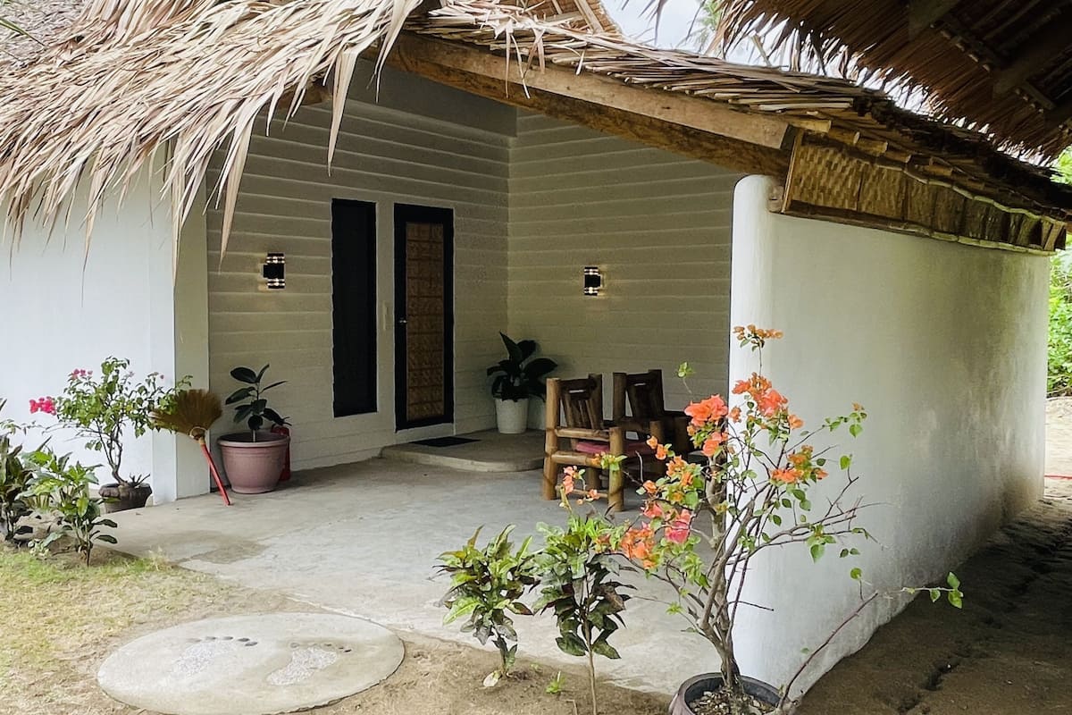 A small, single-story building with a thatched roof, a concrete porch, and potted plants.