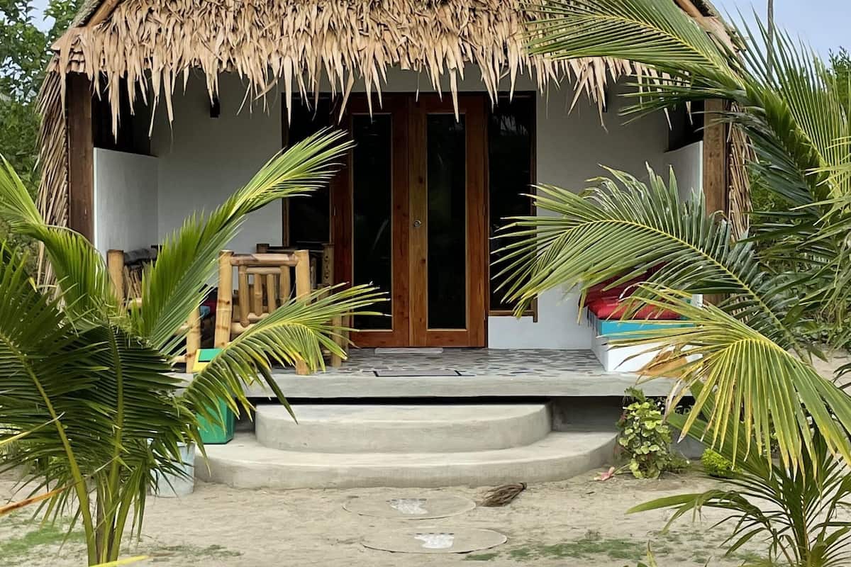 A thatched-roof bungalow with a wooden door and a concrete porch, surrounded by palm trees and a grassy area with stepping stones.