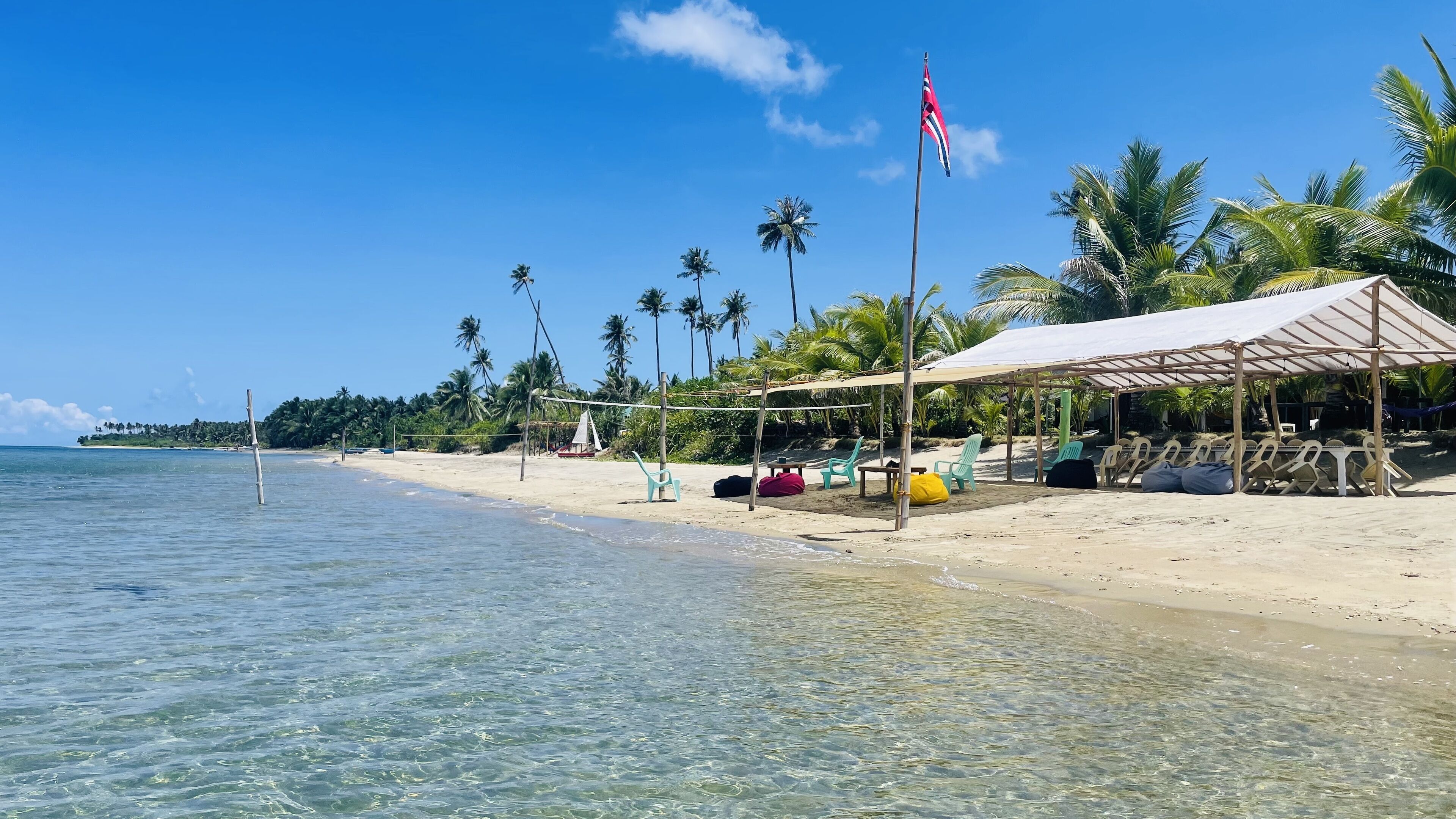 A beach with clear water, a covered seating area with chairs, and a flag.