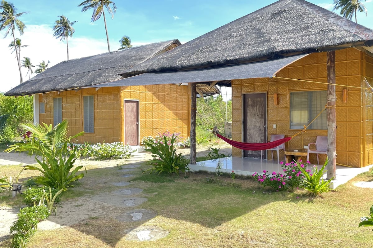 A wooden house with a thatched roof, a hammock, and a small patio area with chairs and a table.