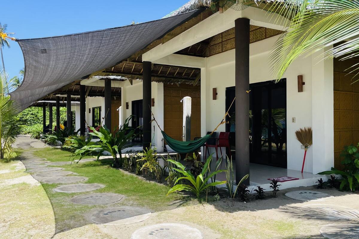 A covered outdoor area with a hammock, palm trees, and a pathway leading to a building.