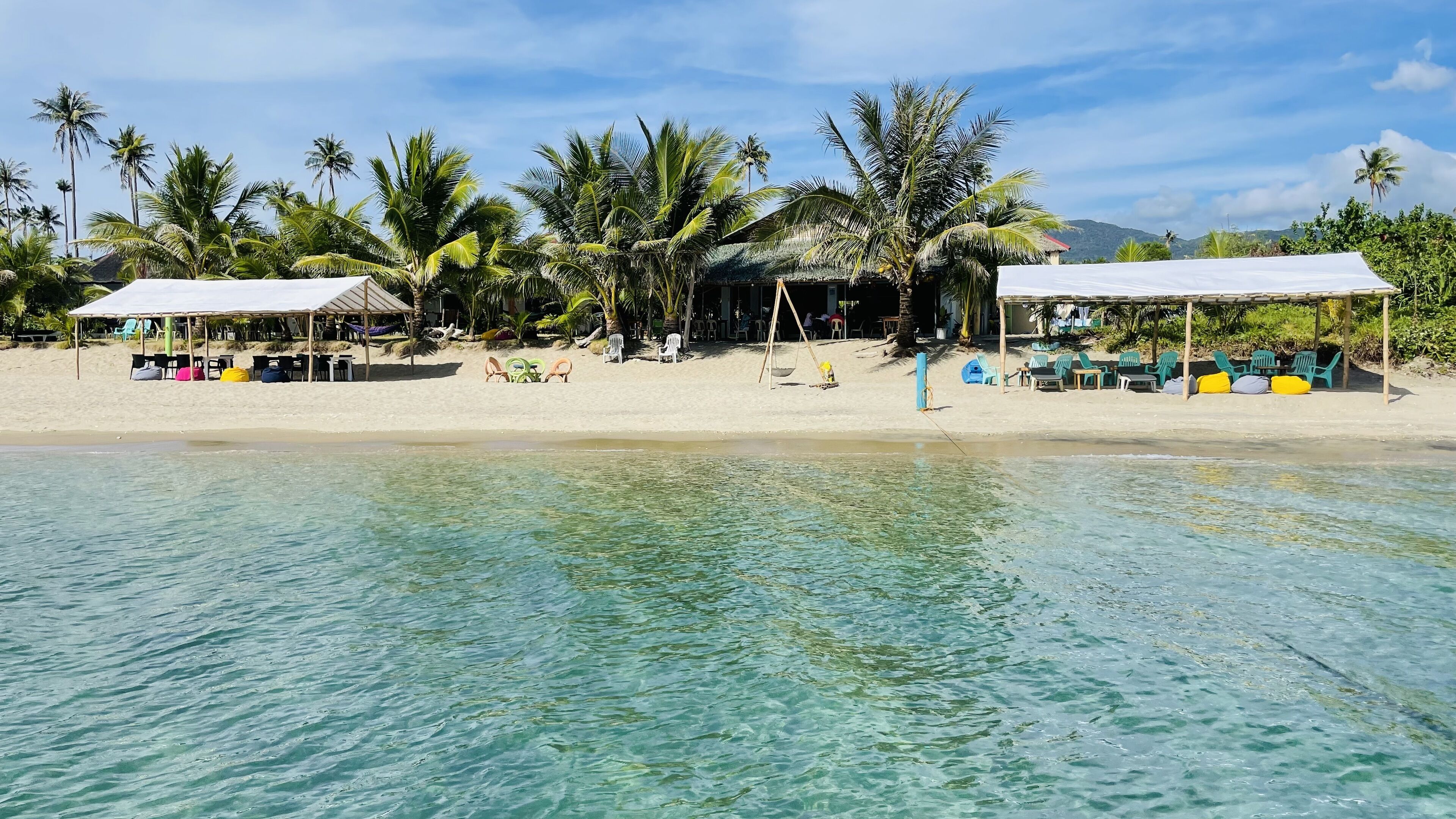 A beach with clear turquoise water, palm trees, and beach chairs under shaded areas.