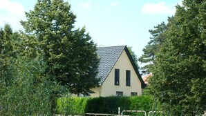 Une maison de plain-pied avec un toit en pente, entourée d’une végétation luxuriante et d’arbres.