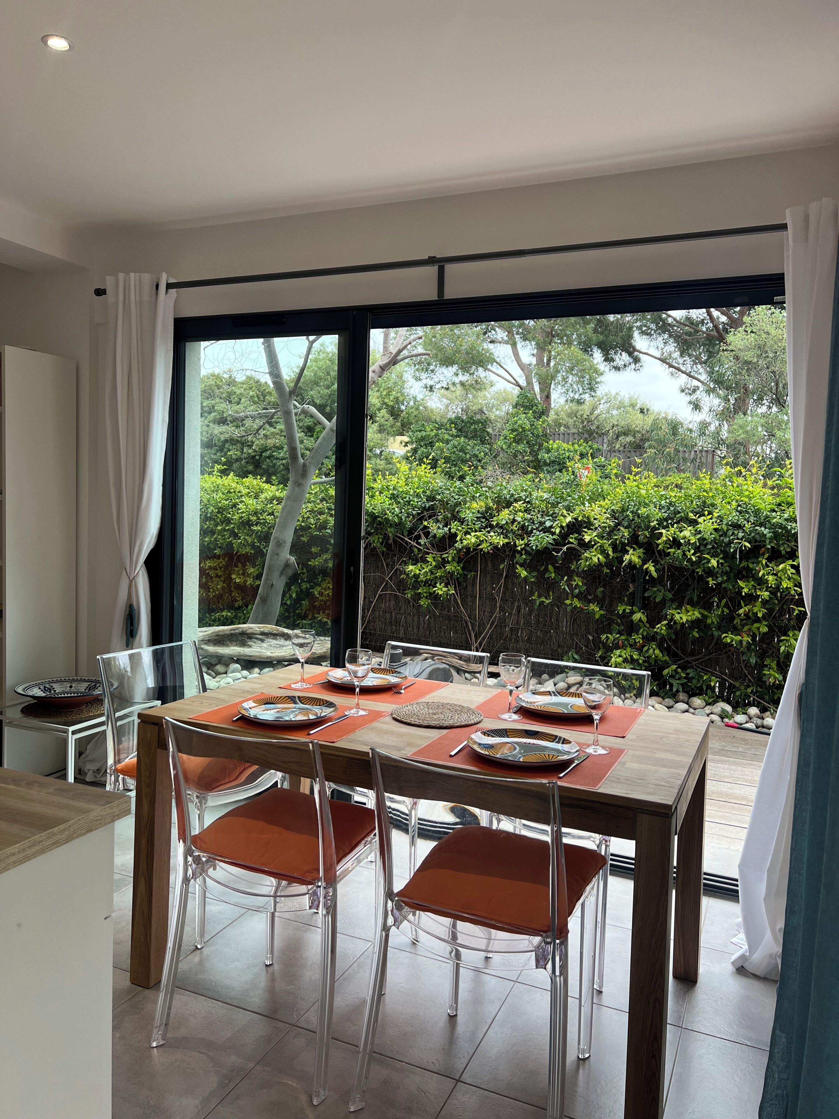Un coin repas avec une table dressée pour six personnes, des chaises transparentes et une vue sur le jardin grâce à de grandes fenêtres.