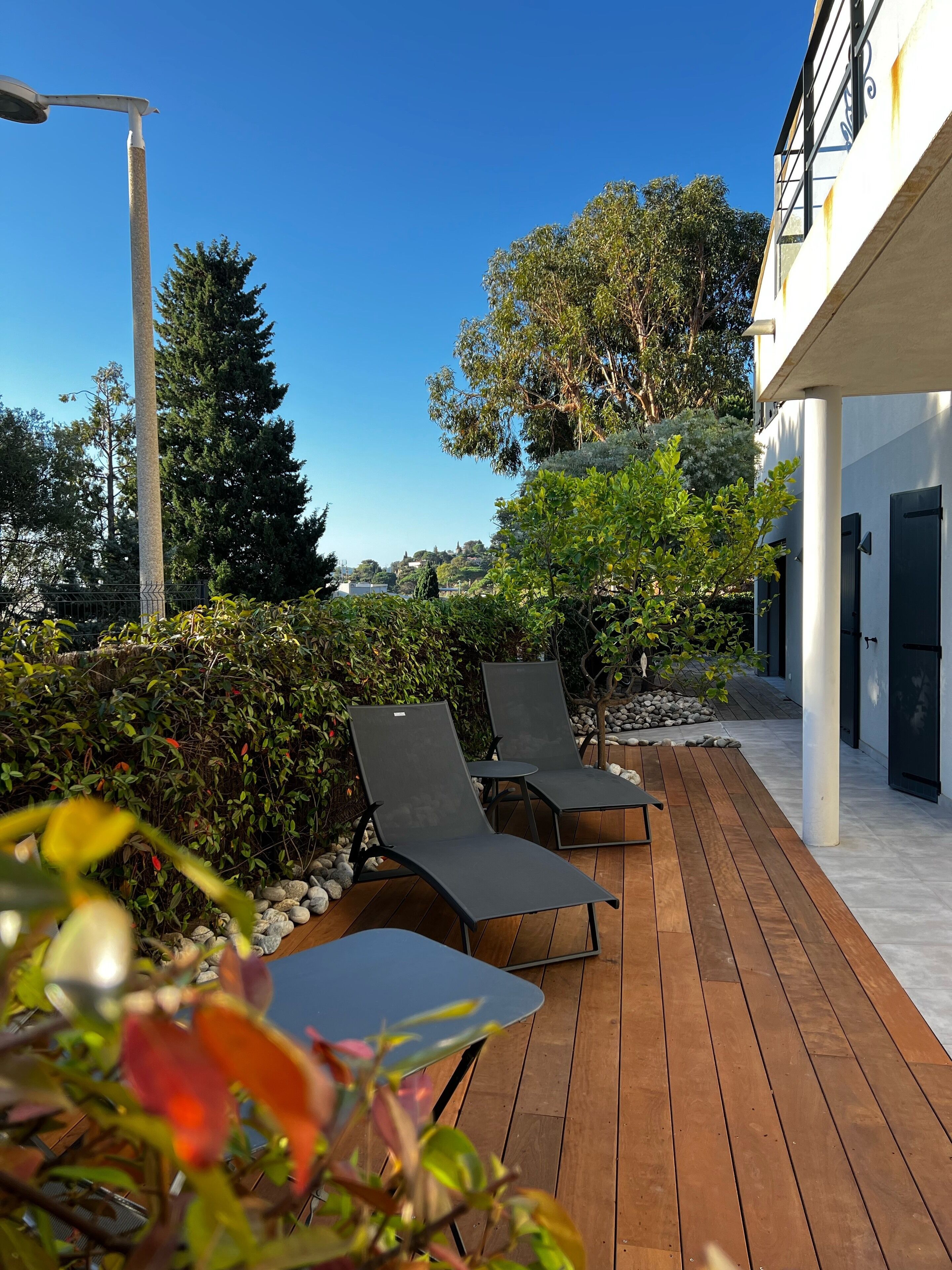 Une terrasse en bois avec des chaises longues, entourée de verdure et bordée d’un bâtiment à la façade blanche.