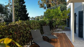 Une terrasse en bois avec des chaises longues, entourée de verdure et bordée d’un bâtiment à la façade blanche.