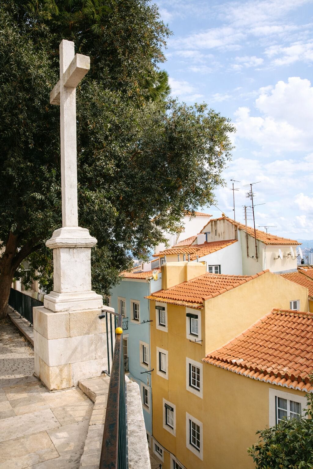 A white cross monument on a stone pedestal, surrounded by buildings with terracotta roofs and a tree.