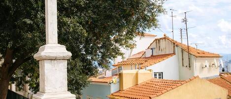 A white cross monument on a stone pedestal, surrounded by buildings with terracotta roofs and a tree.