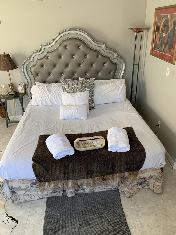 A neatly made bed with white linens and decorative pillows, a bedside table with a lamp and a tissue box, and a small tray with towels and toiletries. - Fantastic Vibrant Art Home<br>King bed/ Wifi/ Sound system<br>Smart TV/ Bike (Richmond)