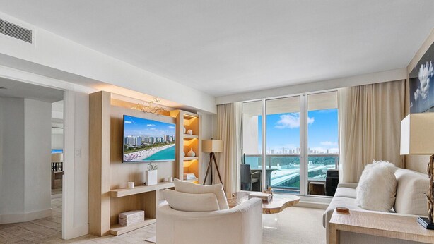 A modern living room with a large TV, comfortable seating, a bookshelf, and a view of a cityscape through large windows. - Luxurious 2/2 CIty View Condo 1145 - Located at 1 Hotel & Homes South Beach (Miami Beach)