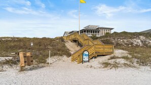Una playa arenosa con una escalera de madera que lleva a un edificio, un asta de bandera y un cartel.