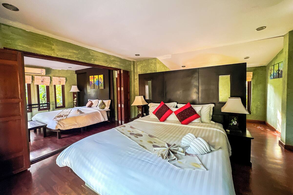 A hotel room with two beds, a large bed with a decorative pillow, a smaller bed, and a nightstand with a lamp.