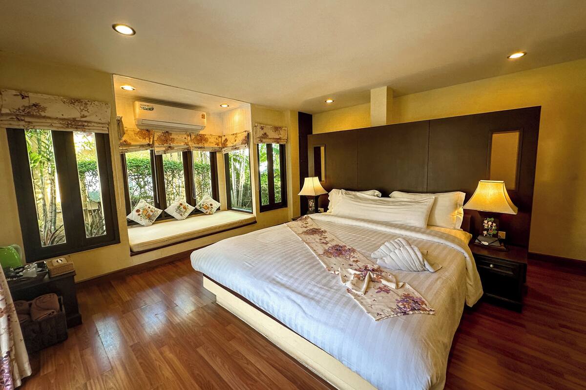 A spacious bedroom with a large bed, two bedside tables with lamps, a window seat with curtains, and a view of greenery outside.