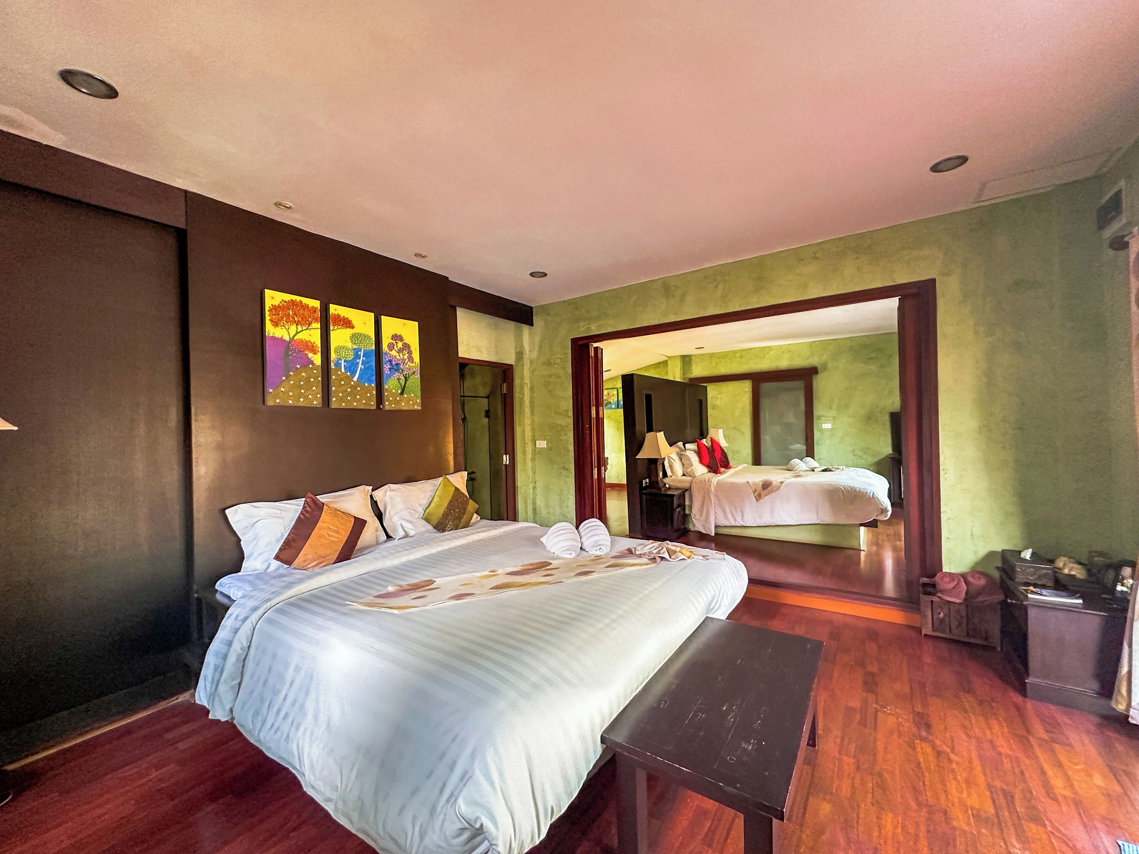 A hotel room with a large bed, bedside tables, a bench, and a view of another room through an open doorway.
