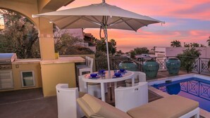 Un espace piscine aménagé sur le toit, offrant une vue sur le coucher de soleil, entouré de palmiers et comprenant un coin repas avec des chaises et une table.