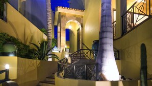 Un escalier bien éclairé, doté d’une rampe et bordé de cactus et de plantes, mène à un bâtiment avec un balcon offrant une vue sur le ciel.