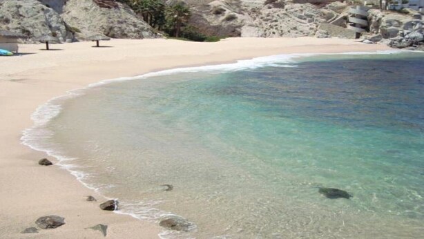 Une plage à l’eau turquoise et limpide, avec un rivage sablonneux et des affleurements rocheux.