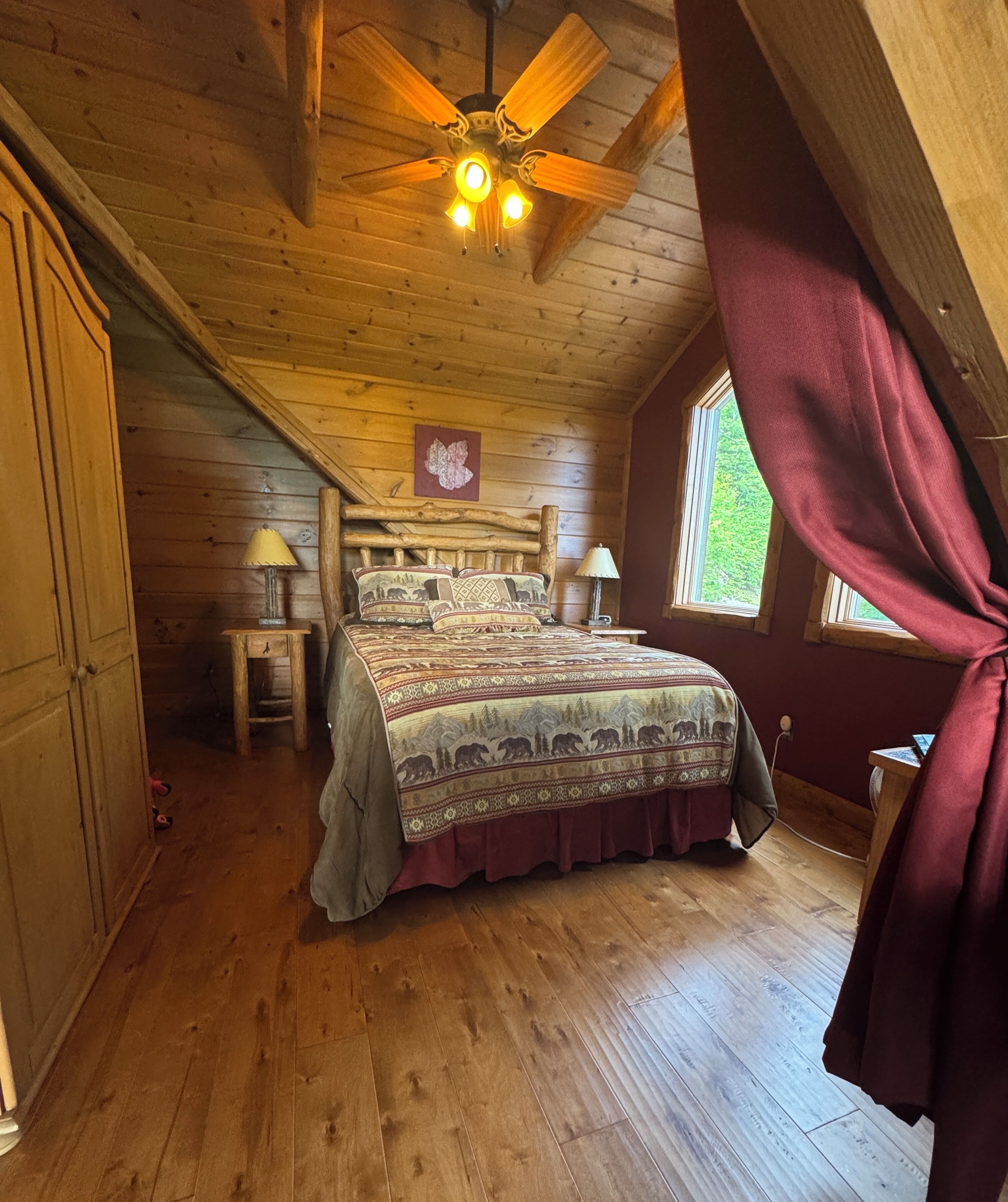 A cozy attic bedroom with a wooden ceiling, a bed with patterned bedding, two bedside lamps, and a window with a view of greenery.