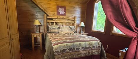 A cozy attic bedroom with a wooden ceiling, a bed with patterned bedding, two bedside lamps, and a window with a view of greenery.