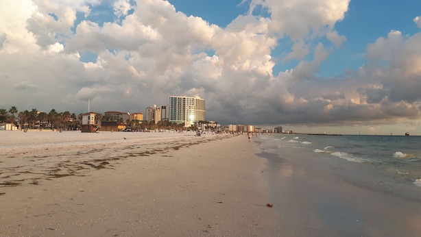 Una playa con un hotel grande al fondo, palmeras y cielo despejado.