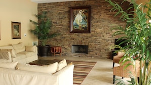 Una sala de estar con chimenea de ladrillo, sofás color beige, alfombra estampada y una planta grande en maceta.