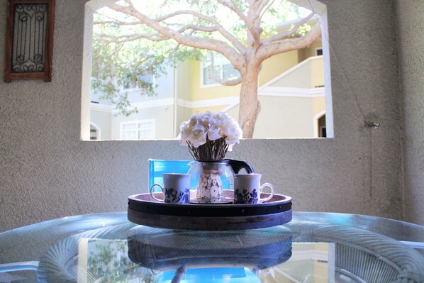A table with a glass top, a tray with two cups and a vase with flowers, and a window with a tree view.