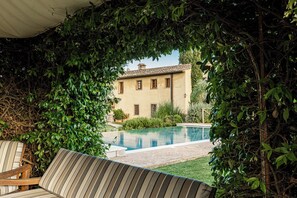 Zona junto a la piscina con un área de descanso cubierta, piscina y un edificio al fondo.