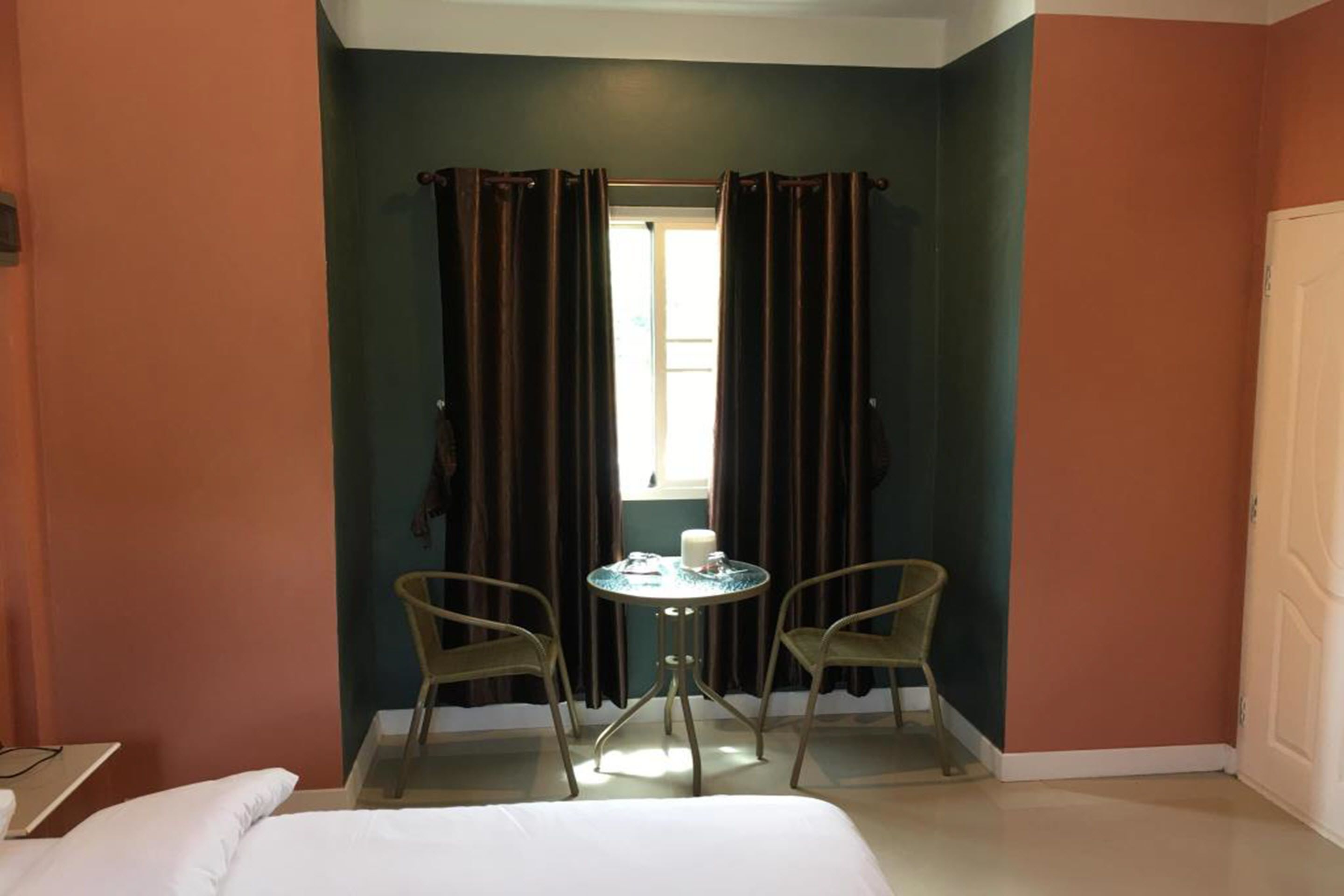 A room with a bed, a small round table with two chairs, a window with curtains, and a door.
