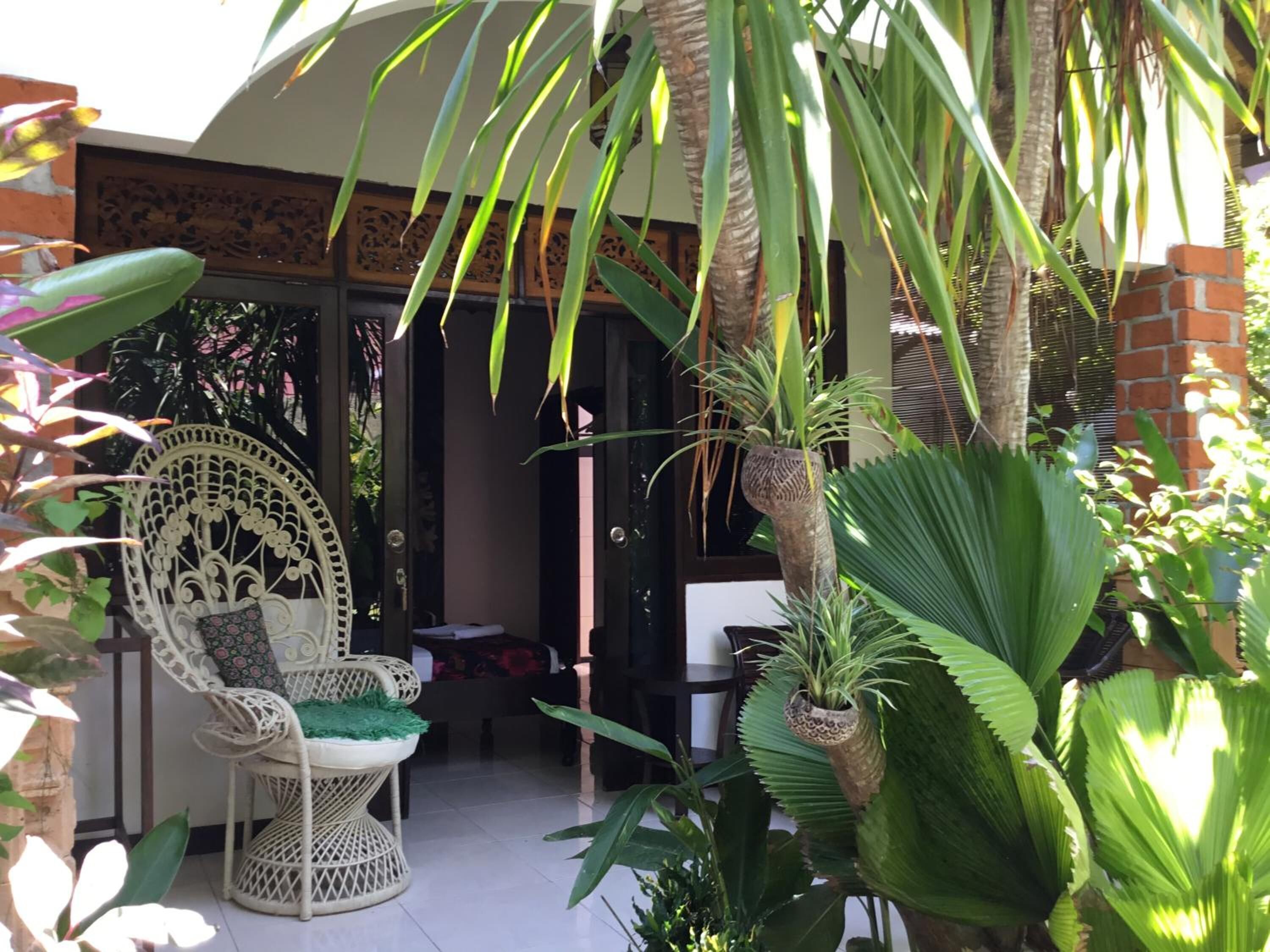 A covered outdoor seating area with wicker furniture, surrounded by various tropical plants and a palm tree.