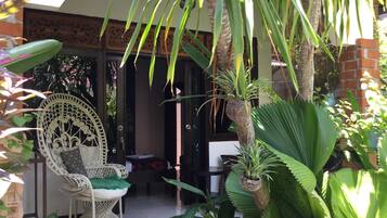 A covered outdoor seating area with wicker furniture, surrounded by various tropical plants and a palm tree.