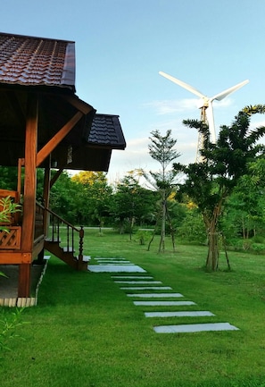 A wooden structure with a staircase leading to a covered area, a grassy pathway, and a wind turbine in the background. - Rai Lung Tui Homestay (Prachuap Khiri Khan)