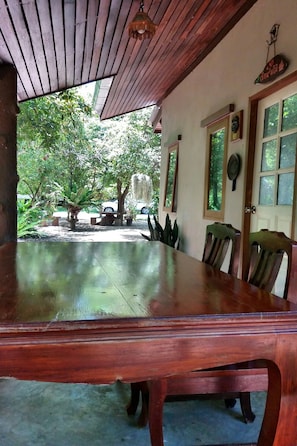 A wooden dining table with chairs, a wooden ceiling, and a view of trees and a car outside. - Rai Lung Tui Homestay (Prachuap Khiri Khan)