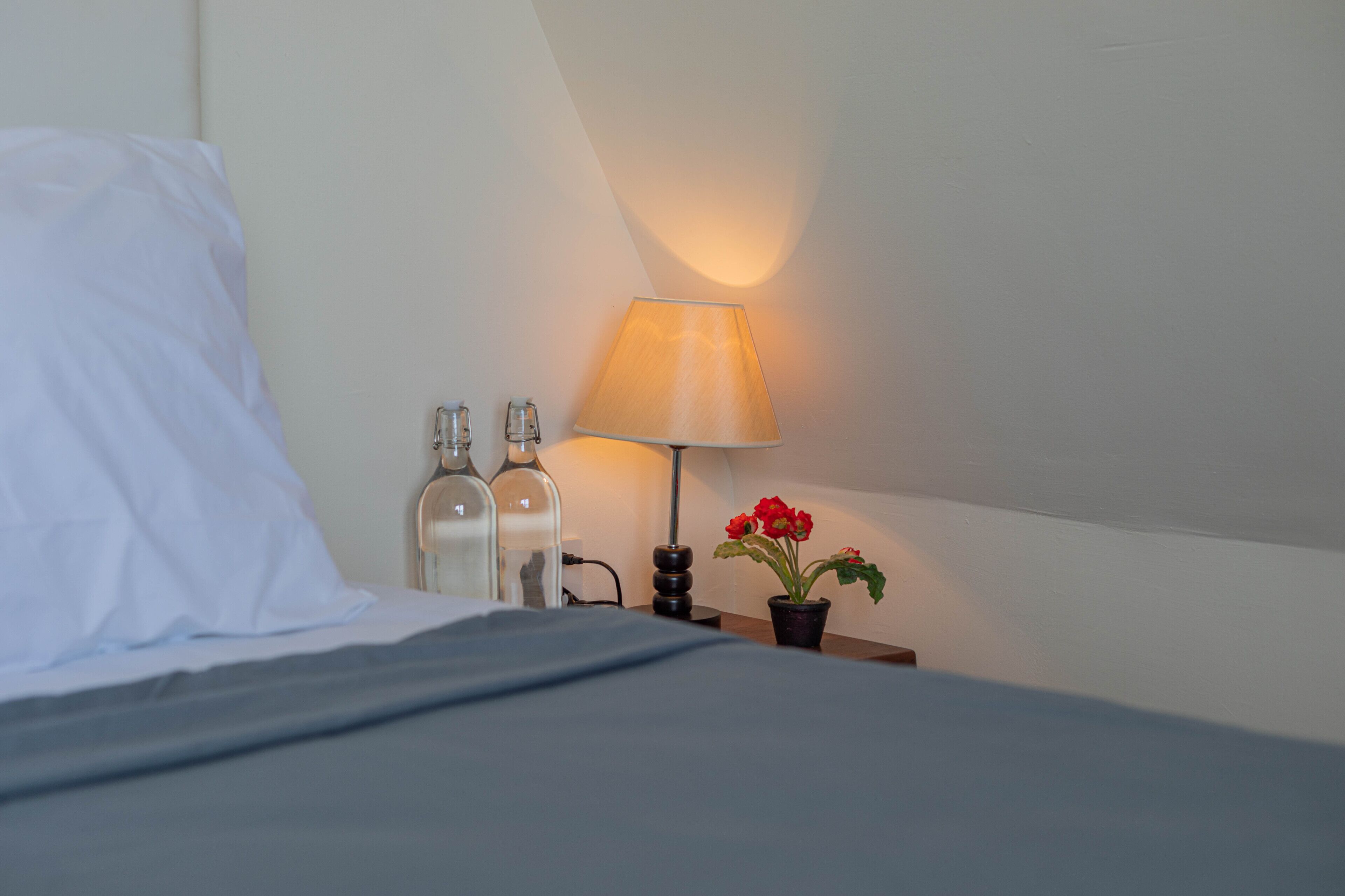 A neatly made bed with white linens, a bedside table with a lamp, two bottles, and a potted plant with red flowers.