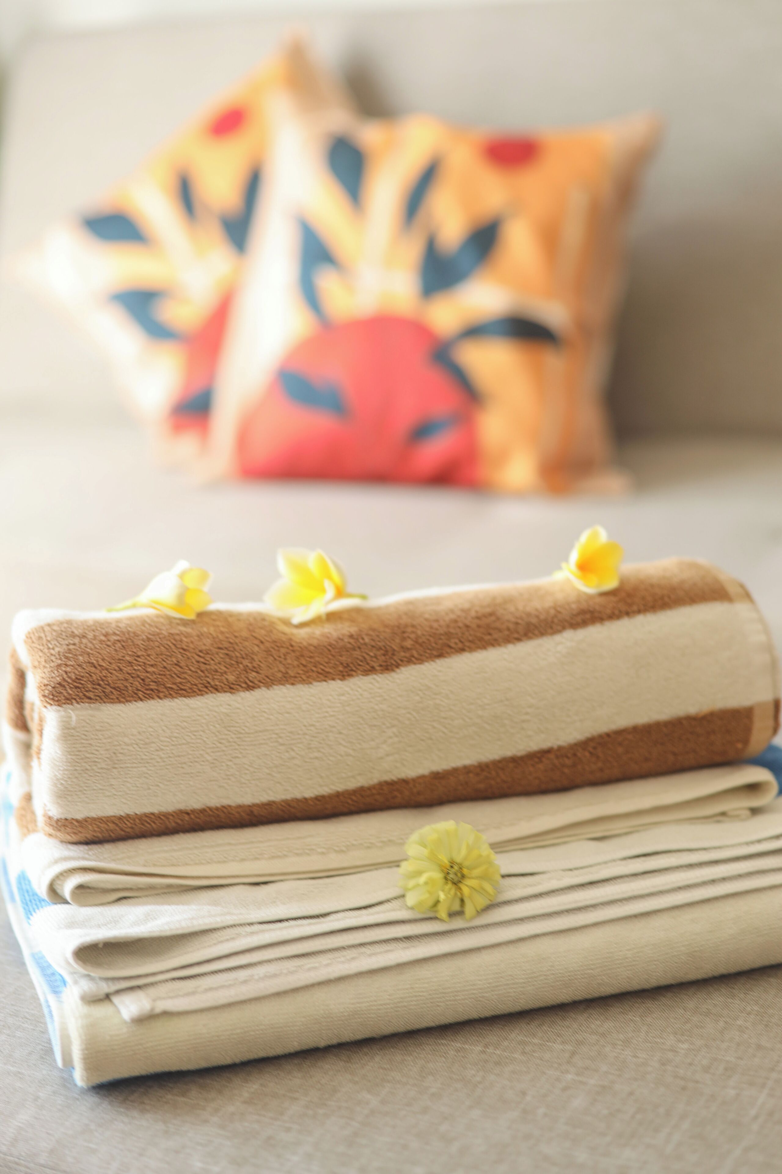 Toalha dobrada com uma flor decorativa, colocada sobre uma cama com um travesseiro colorido ao fundo.