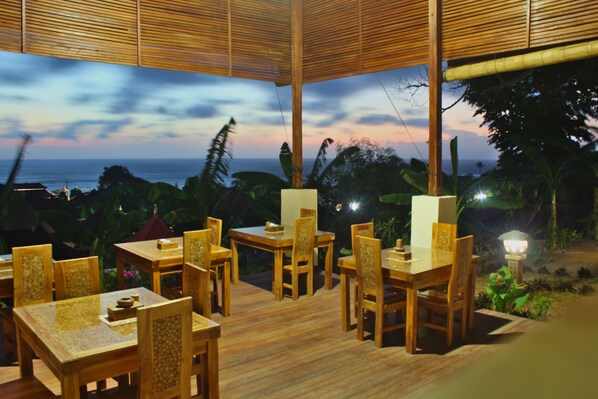 An outdoor dining area with wooden tables and chairs, surrounded by lush greenery and overlooking a scenic view of the ocean at dusk.