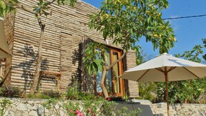 Un edificio rústico de madera con techo de paja, rodeado de exuberante vegetación y un muro de piedra.