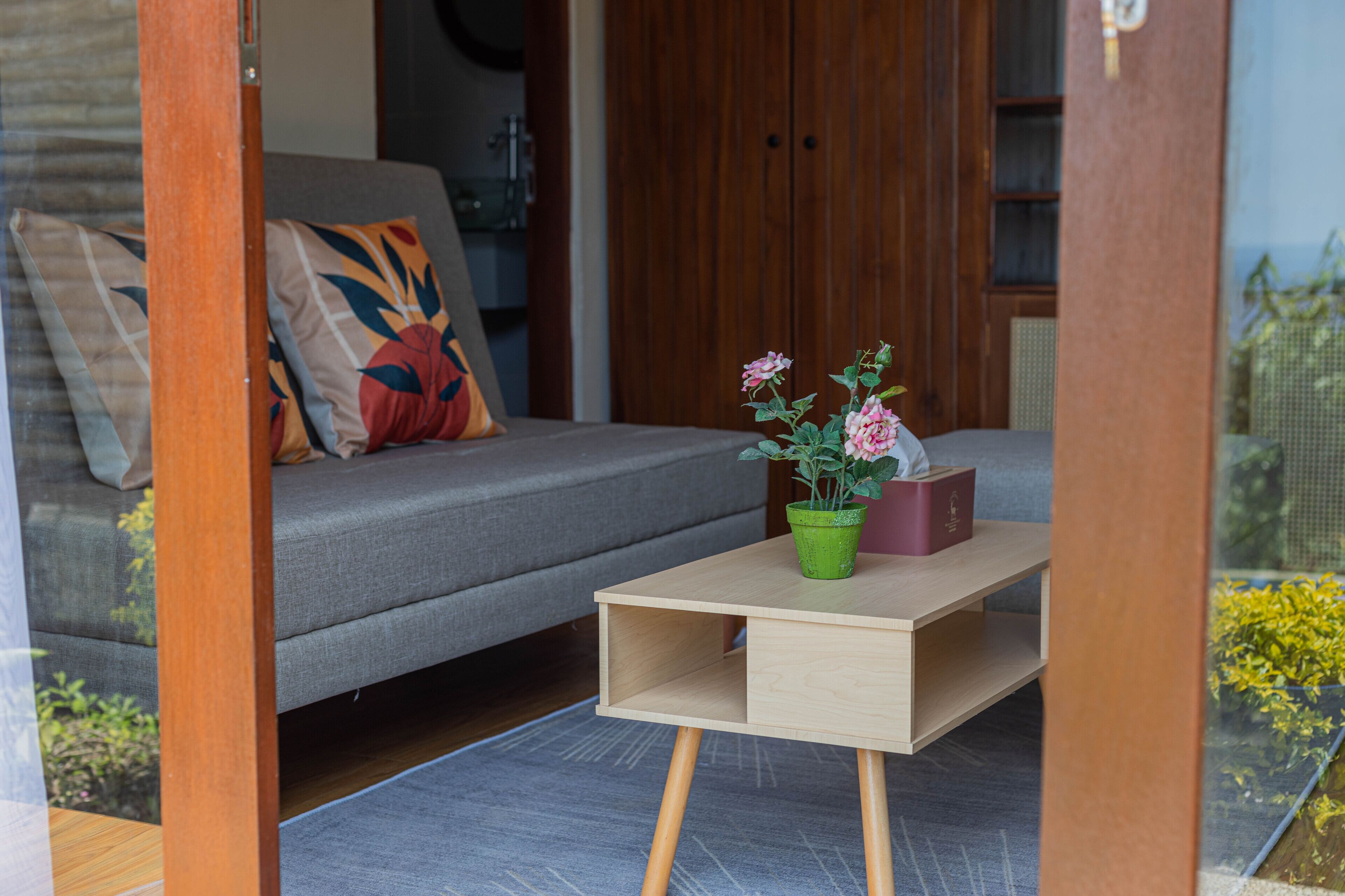 Una sala de estar moderna con un sofá gris, una mesa de centro de madera con una maceta verde y un libro rojo, y una puerta de madera al fondo.