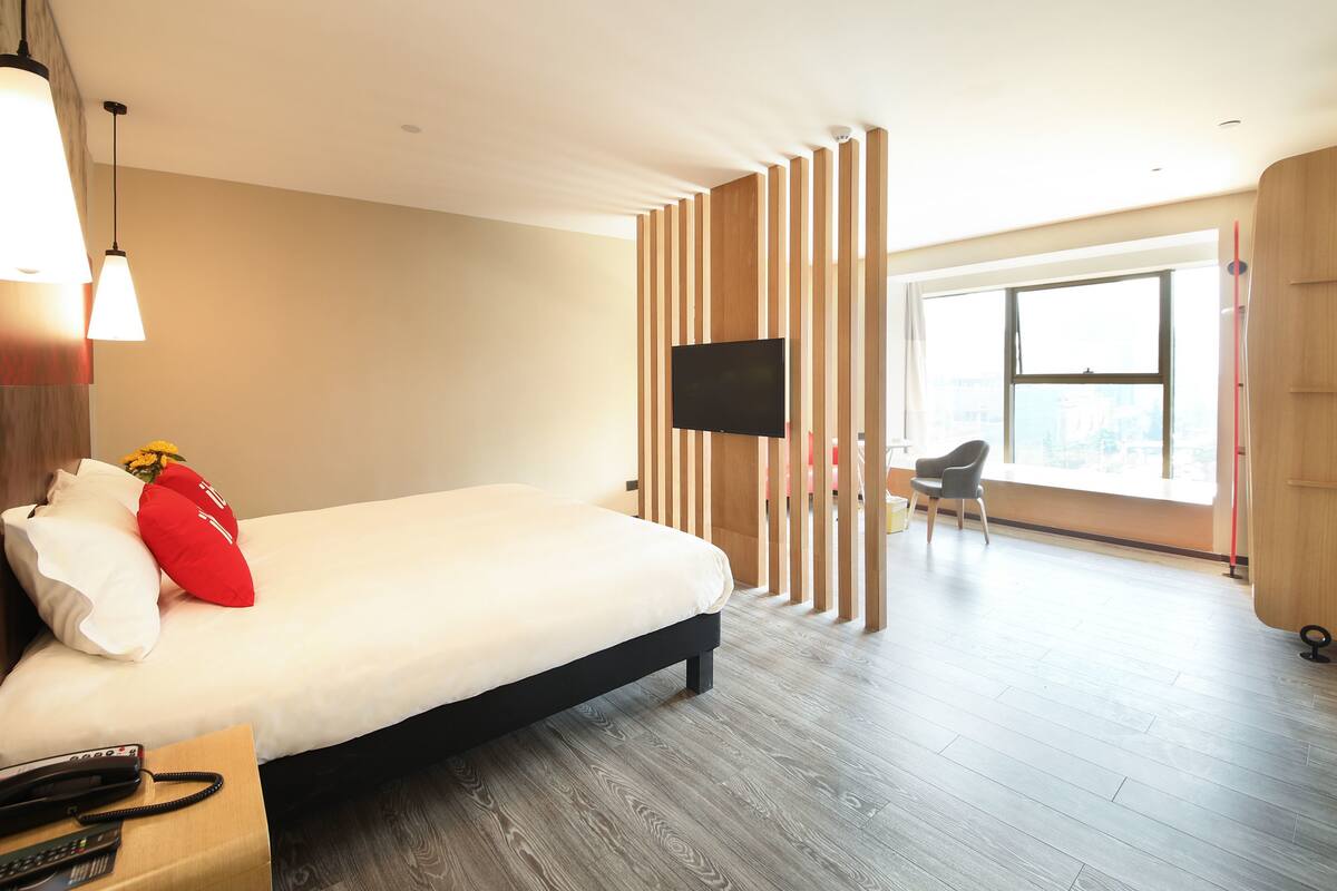 Habitación de hotel moderna con una cama grande, un televisor de pantalla plana montado en un panel de madera, piso de madera y un ventanal con vistas a la ciudad.