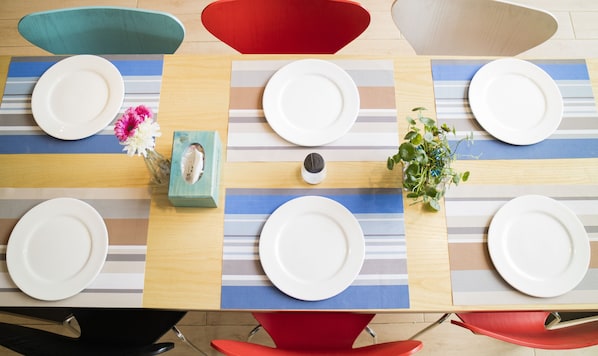 Une table à manger avec quatre assiettes blanches, un salier, un petit vase avec des fleurs et une boîte. La table est dressée avec des sets de table à rayures et entourée de chaises colorées.