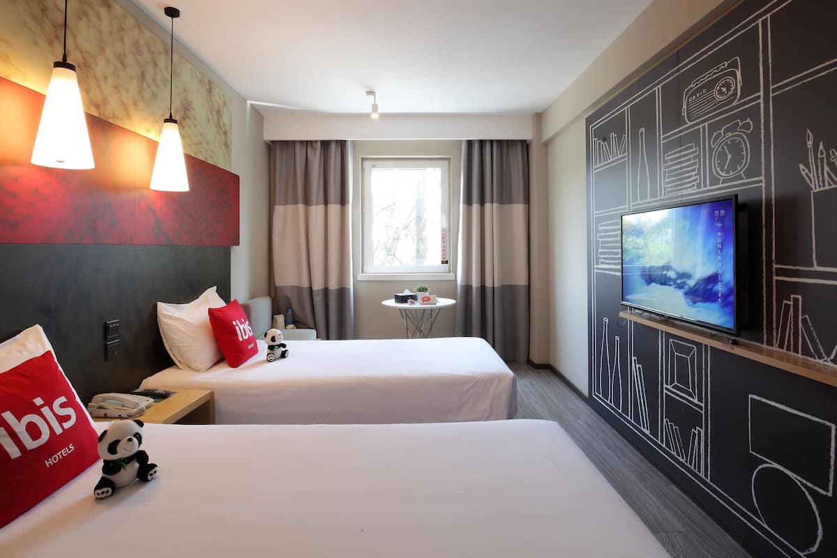 Habitación de hotel con dos camas, televisión, una mesita y ventana con cortinas.