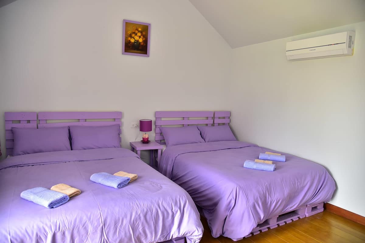 Une chambre avec deux lits, chacun agrémenté de draps et d’oreillers violets, une petite table de chevet avec une lampe et un climatiseur fixé au mur.