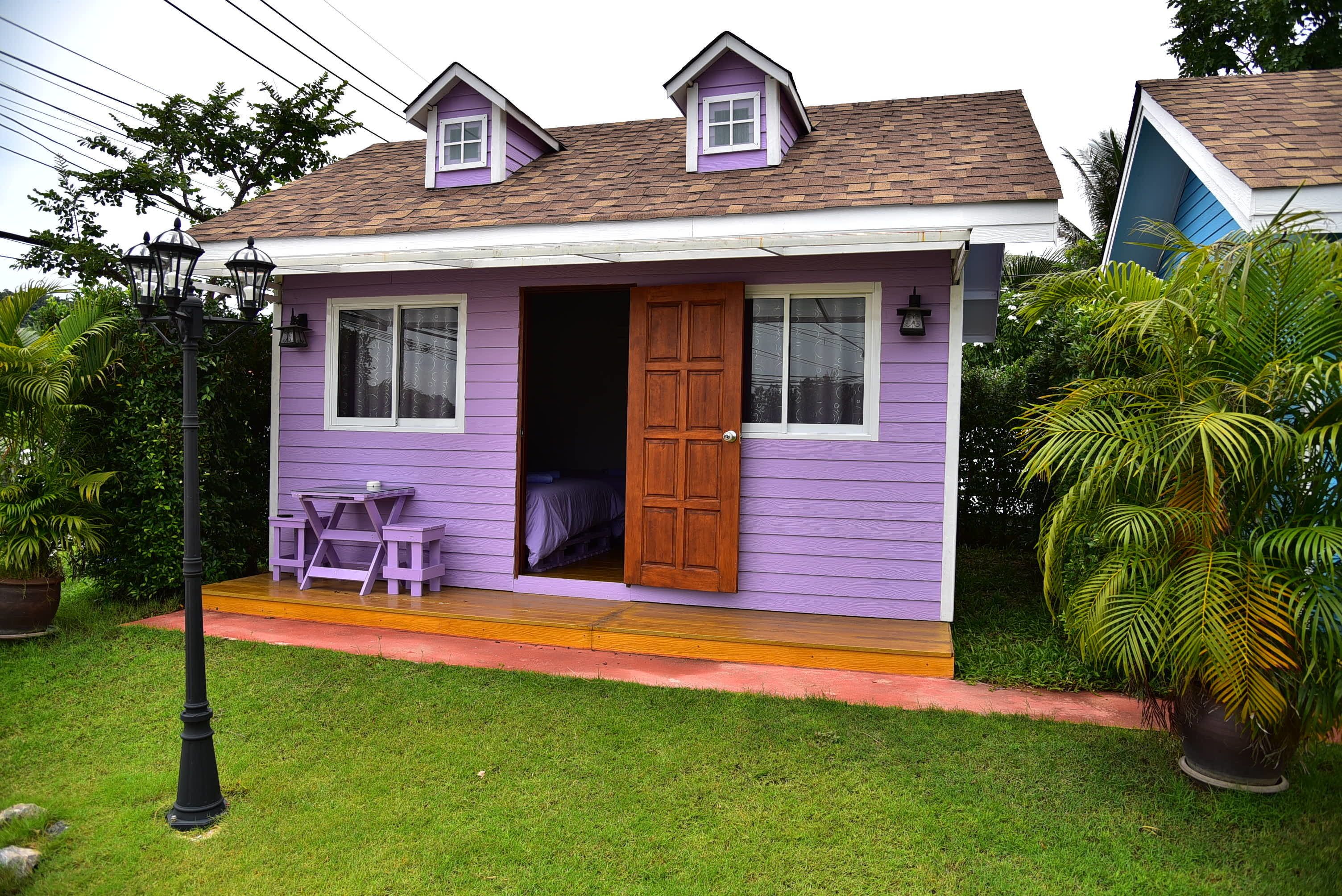 A small, single-story house with a purple exterior, a wooden door, and a small porch with two chairs.
