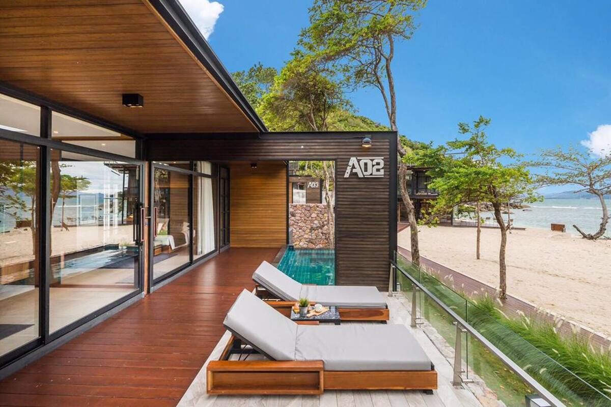A modern beachside villa with a glass-enclosed balcony, a pool, and lounge chairs.
