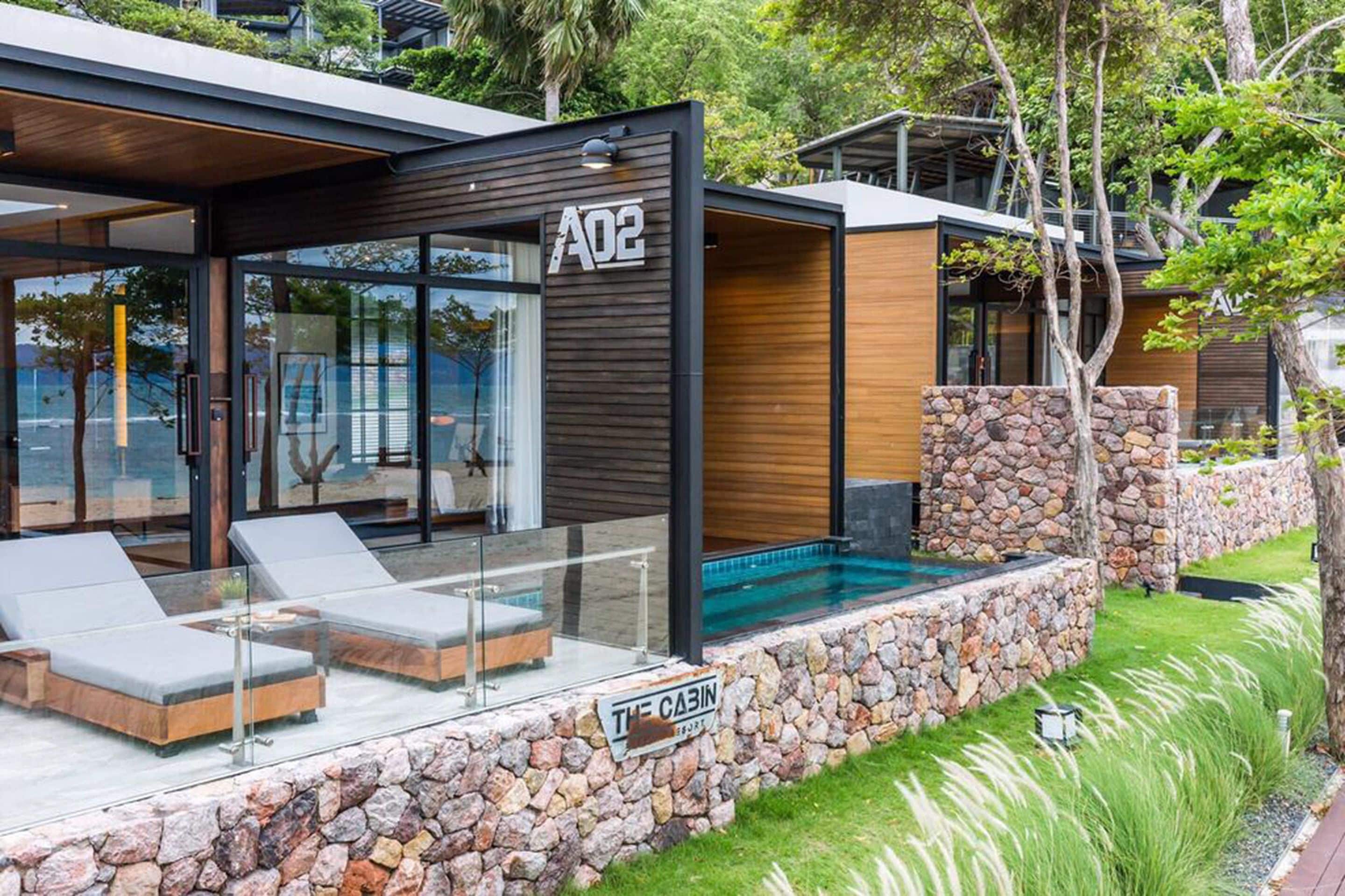 A modern cabin with a glass exterior, a stone wall, and a pool area.