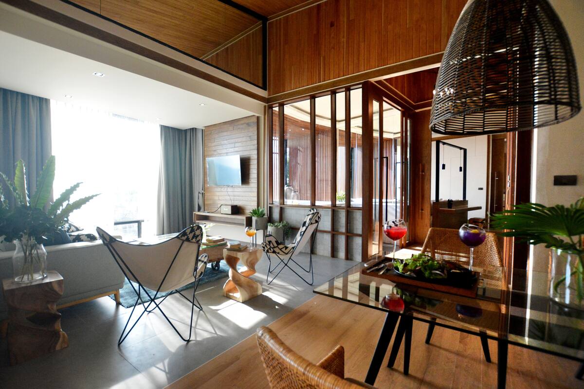 A modern living room with a glass coffee table, wooden flooring, and a large window with curtains. There are several chairs and a sofa, and a hanging light fixture.
