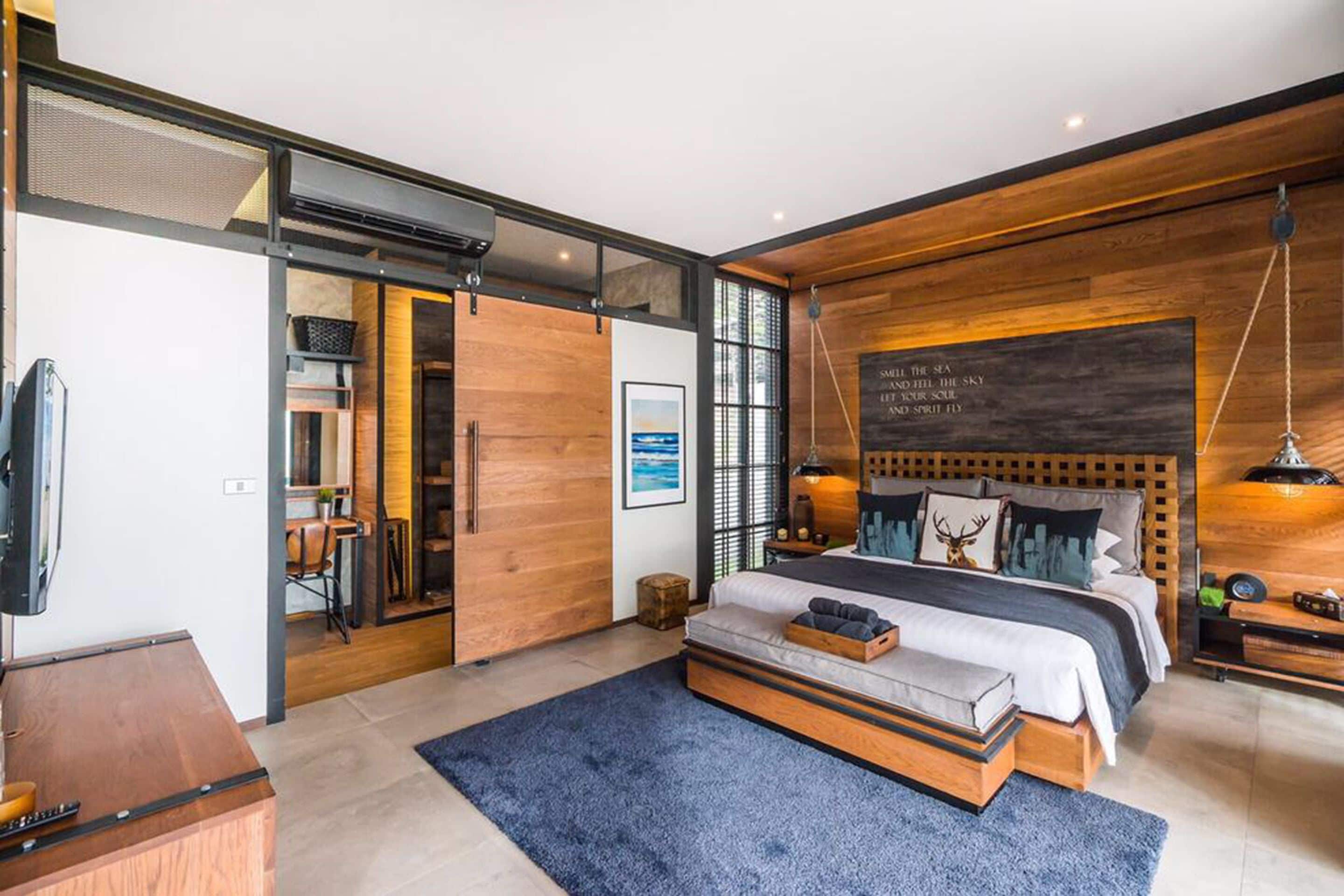 A modern bedroom with a large bed, wooden accents, a mounted TV, and a view of the sea.