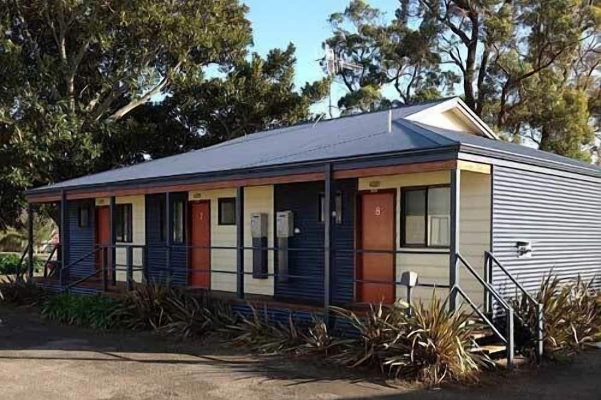 A single-story motel with a flat roof, a covered porch, and a small front yard with plants.